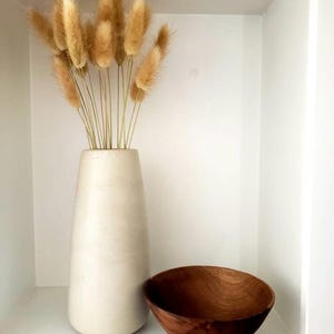 May include: A neutral-toned interior decor arrangement featuring a tall, off-white vase holding dried, fluffy, tan-coloured bunny tail grass. A small, round, wooden bowl sits beside the vase. The items are displayed in a white shelf.