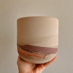 May include: A cylindrical ceramic planter featuring a matte, light beige upper section and a textured lower section with layered, horizontal bands of earthy terracotta, deep maroon, and soft tan colors, creating a natural, stratified appearance.