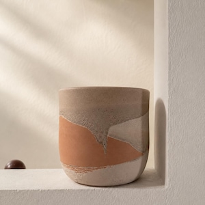 Peut inclure: Un pot de fleurs cylindrique en céramique présentant une finition texturée et multicolore avec des tons terreux de terre cuite, beige et blanc cassé. La surface affiche un motif abstrait en couches avec des détails granulaires visibles.