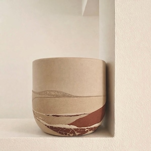 May include: A cylindrical beige ceramic pot with a textured landscape design in shades of tan and rust. The pot has a smooth upper section and a textured lower section, creating a layered effect. The pot is displayed on a white shelf.