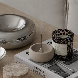 Peut inclure: Nature morte avec un bol argenté, un petit porte-encens beige avec un bâtonnet allumé, une bougie à imprimé léopard et des dessous de verre en marbre. Un livre intitulé "Bauhaus Alvar Aalto" est également présent.
