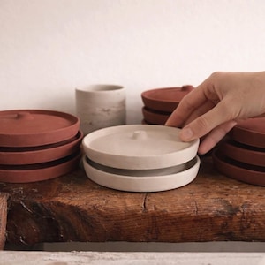 Peut inclure: Plusieurs assiettes et couvercles en céramique couleur terre cuite sont empilés. Une main interagit avec les assiettes. Les assiettes sont posées sur une surface en bois rustique, avec un fond neutre. Les assiettes sont probablement pour servir ou ranger, environ 15 cm de diamètre.