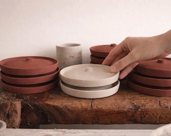 May include: Terracotta-coloured ceramic plates and lids are stacked in sets. A hand interacts with the plates. The plates sit on a rustic wooden surface, with a neutral background. The plates are likely for serving or storage, with a diameter of approximately 15 cm.