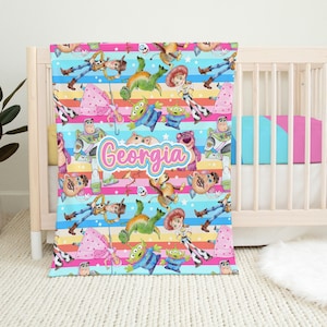 Peut inclure: Une couverture pour b&eacute;b&eacute; color&eacute;e avec un motif r&eacute;p&eacute;t&eacute; de personnages de la franchise Toy Story. La couverture est bleue avec des rayures roses, jaunes et vertes. Les personnages incluent Woody, Buzz l'&Eacute;clair, Jessie et les extraterrestres. Le nom "Georgia" est &eacute;crit en lettres roses au centre de la couverture.