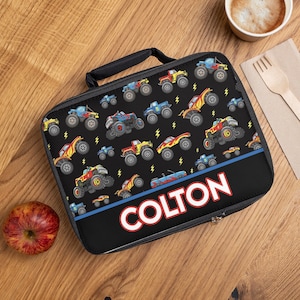 May include: Black lunch bag with a zipper closure and a handle. The bag has a pattern of colorful monster trucks on a black background. The name "COLTON" is printed in white letters on a red and blue stripe.