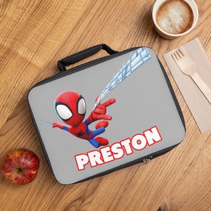 May include: A gray lunch bag with a black zipper and handle. The bag features a cartoon image of Spiderman swinging on a web with the name Preston printed in red letters.