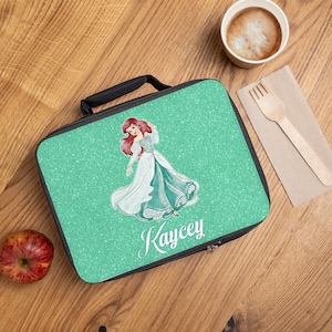 May include: A green lunch bag with a glitter finish featuring a cartoon image of Ariel from The Little Mermaid. The bag has a black zipper and handle and the name "Kaycey" is printed on the front.
