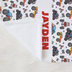 May include: White fleece blanket with a red and blue monster truck pattern. The name "JAYDEN" is printed in red on the right side of the blanket.