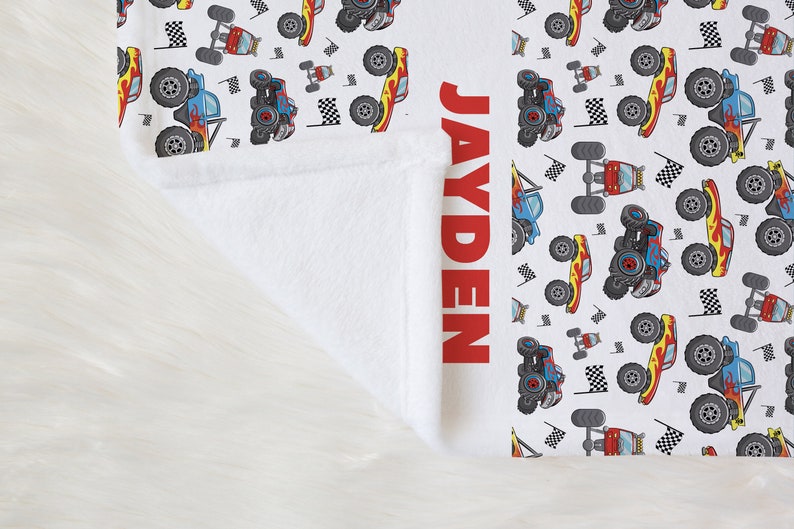 May include: White fleece blanket with a red and blue monster truck pattern. The name "JAYDEN" is printed in red on the right side of the blanket.