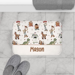 May include: White bath mat with a brown border featuring a repeating pattern of Toy Story characters, including Woody, Buzz Lightyear, and Mr. Potato Head. The mat has the name "Mason" printed in brown letters.