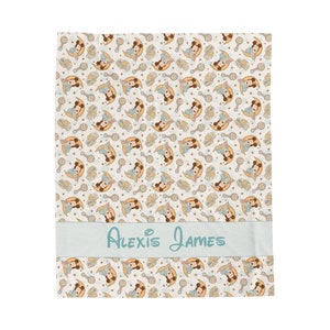 May include: A white baby blanket with a light blue border and a repeating pattern of a cartoon mouse holding a rattle. The blanket has a light blue personalized name tag that reads "Alexis James".