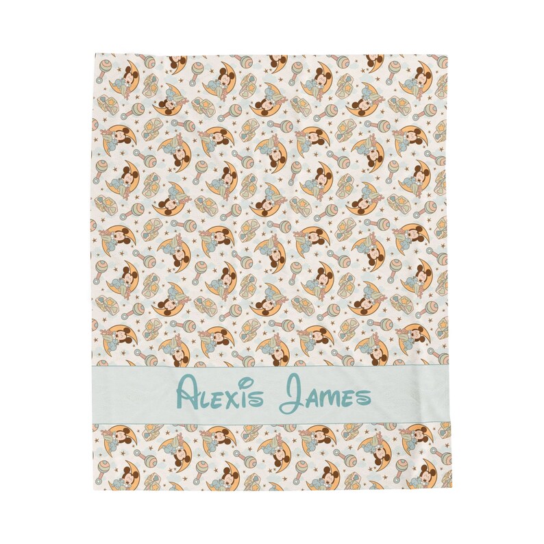 May include: A white baby blanket with a light blue border and a repeating pattern of a cartoon mouse holding a rattle. The blanket has a light blue personalized name tag that reads "Alexis James".