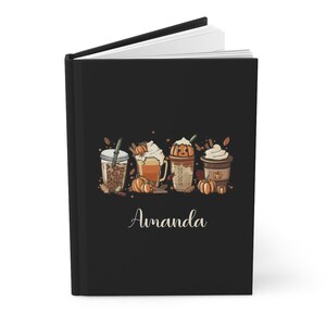K&ouml;nnte beinhalten: Schwarzes Journal mit dem personalisierten Namen "Amanda" und einer Grafik von vier Herbst-Kaffeegetr&auml;nken mit K&uuml;rbisgew&uuml;rz, Schlagsahne und Herbstbl&auml;ttern.