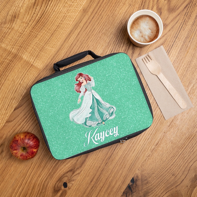 May include: A green lunch bag with a glitter finish featuring a cartoon image of Ariel from The Little Mermaid. The bag has a black zipper and handle and the name "Kaycey" is printed on the front.