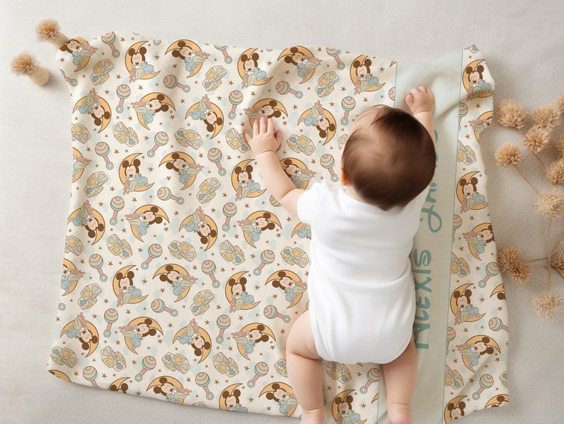 May include: A cream-colored baby blanket with a repeating pattern of Mickey Mouse characters in blue and yellow. The blanket has a green border with the name "Alexis" written vertically on the side.