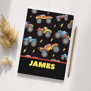 May include: Black notebook with a monster truck design. The notebook has a red stripe and the name "JAMES" printed on the front.