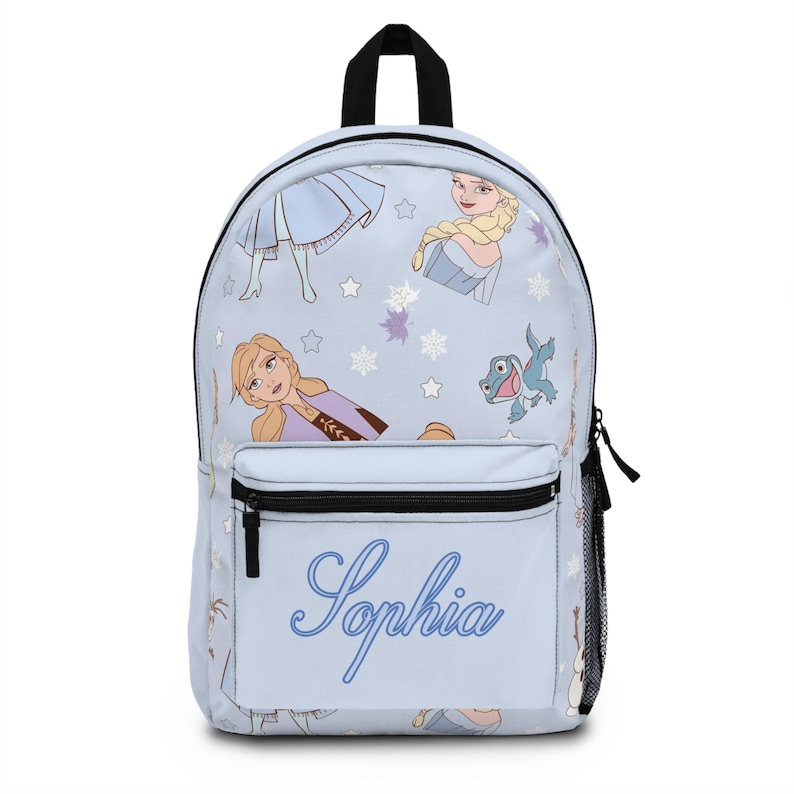 Puede incluir: Una mochila azul claro con ilustraciones de personajes y copos de nieve. Tiene un asa y cremallera negras, con el nombre "Sophia" en escritura azul en la parte delantera. Un bolsillo de malla est&aacute; en el lateral.