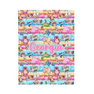 Peut inclure: Une couverture color&eacute;e avec un motif r&eacute;p&eacute;t&eacute; de personnages de Toy Story sur un fond ray&eacute; arc-en-ciel. Le nom "Georgia" est &eacute;crit en lettres roses au centre de la couverture.