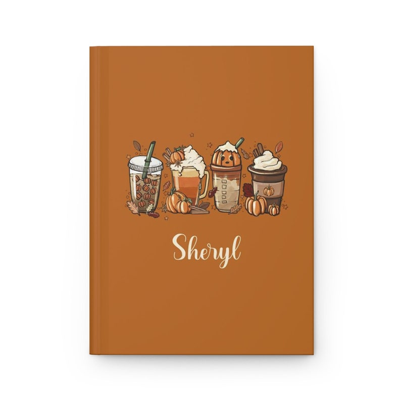 K&ouml;nnte beinhalten: Ein braunes Notizbuch mit einem herbstlichen Design, das vier K&uuml;rbisgew&uuml;rz-Lattes mit Schlagsahne und Herbstbl&auml;ttern zeigt. Der Name "Sheryl" ist unten auf dem Notizbuch in wei&szlig;er Kursivschrift geschrieben.