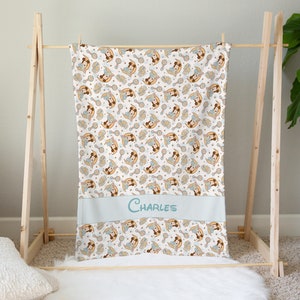 May include: A white baby blanket with a repeating pattern of a cartoon girl holding a rattle. The blanket has a light blue border with the name "CHARLES" embroidered in the center.