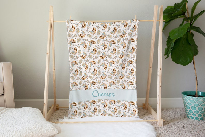May include: A white baby blanket with a repeating pattern of a cartoon girl holding a rattle. The blanket has a light blue border with the name "CHARLES" embroidered in the center.