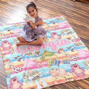 Peut inclure: Une couverture en polaire color&eacute;e sur le th&egrave;me de Toy Story. La couverture pr&eacute;sente une vari&eacute;t&eacute; de personnages du film, notamment Woody, Buzz l'&Eacute;clair, Jessie et Rex. La couverture est d&eacute;cor&eacute;e de rayures, d'&eacute;toiles et du nom "Georgia" en lettres roses.