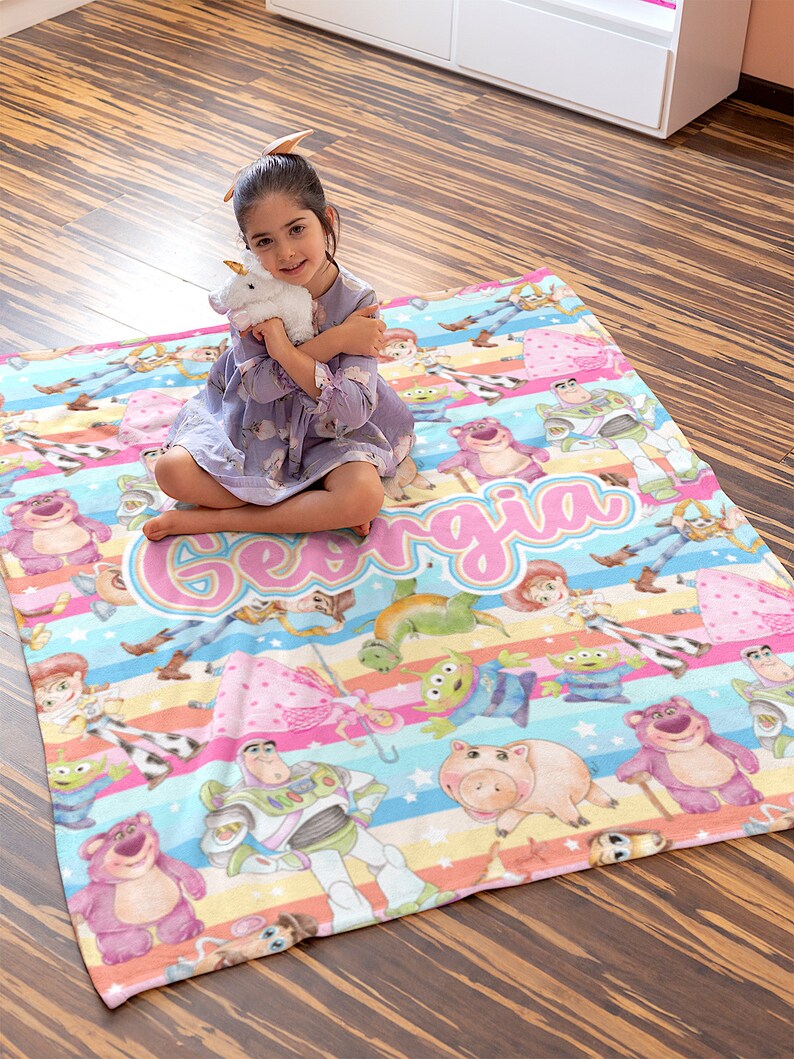 Peut inclure: Une couverture en polaire color&eacute;e sur le th&egrave;me de Toy Story. La couverture pr&eacute;sente une vari&eacute;t&eacute; de personnages du film, notamment Woody, Buzz l'&Eacute;clair, Jessie et Rex. La couverture est d&eacute;cor&eacute;e de rayures, d'&eacute;toiles et du nom "Georgia" en lettres roses.