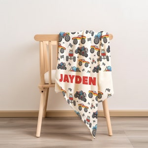 May include: A white and cream colored blanket with a pattern of colorful monster trucks and checkered flags. The blanket is folded and draped over a wooden chair. The name "JAYDEN" is printed in red letters on the blanket.