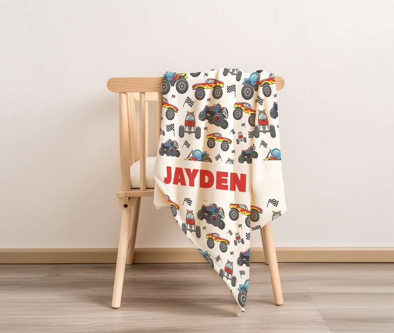 May include: A white and cream colored blanket with a pattern of colorful monster trucks and checkered flags. The blanket is folded and draped over a wooden chair. The name "JAYDEN" is printed in red letters on the blanket.