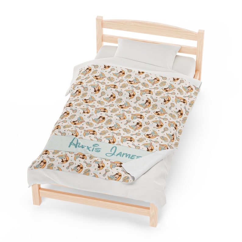 May include: A white and blue personalized baby blanket with a pattern of cartoon characters holding rattles and moons. The blanket has the names "Alexis James" written on it in a light blue font.