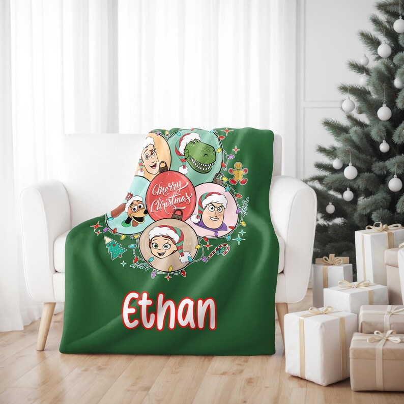 May include: A green Christmas throw blanket featuring cartoon characters in ornaments, with the text "Merry Christmas" and the name "Ethan" in red. The blanket is draped over a white armchair, with a Christmas tree in the background.