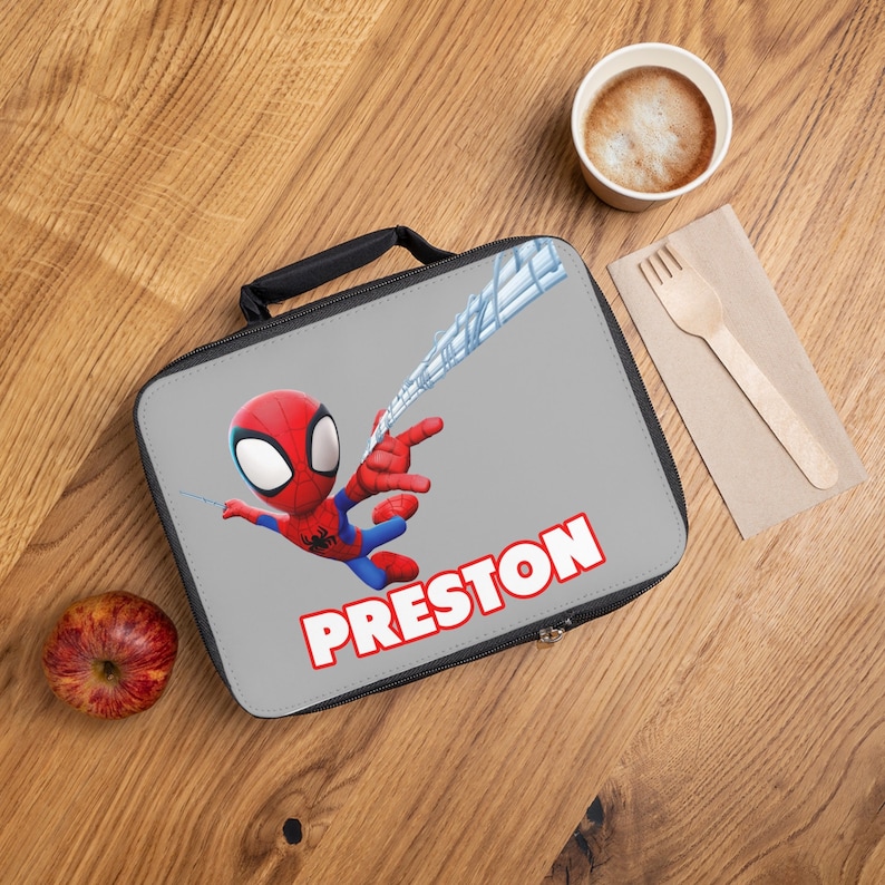 May include: A gray lunch bag with a black zipper and handle. The bag features a cartoon image of Spiderman swinging on a web with the name Preston printed in red letters.