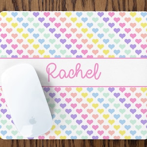 May include: A personalized mouse pad with a white background and a pattern of pastel pink, yellow, blue, and purple hearts. The name "Rachel" is printed in pink cursive on a white banner.