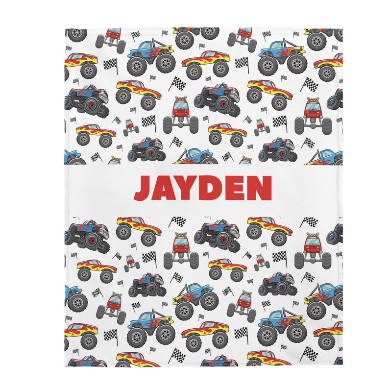 May include: White blanket with a repeating pattern of red, blue, and yellow monster trucks with flames. The name "JAYDEN" is printed in red letters in the center of the blanket.