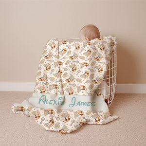 May include: A white baby blanket with a repeating pattern of a cartoon baby holding a rattle and a crescent moon. The blanket has the names "Alexis James" printed on it in a light blue font.