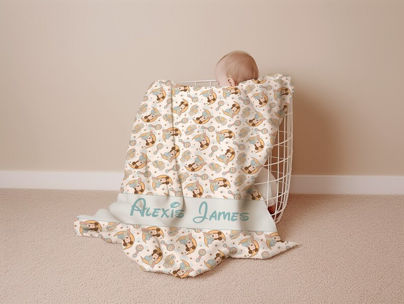 May include: A white baby blanket with a repeating pattern of a cartoon baby holding a rattle and a crescent moon. The blanket has the names "Alexis James" printed on it in a light blue font.