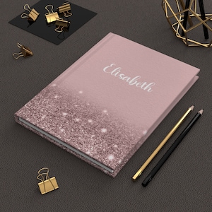 May include: A pink journal with the name "Elisabeth" in silver script. The bottom half of the journal cover is covered in glitter. Gold and black pencils and gold binder clips are next to the journal.