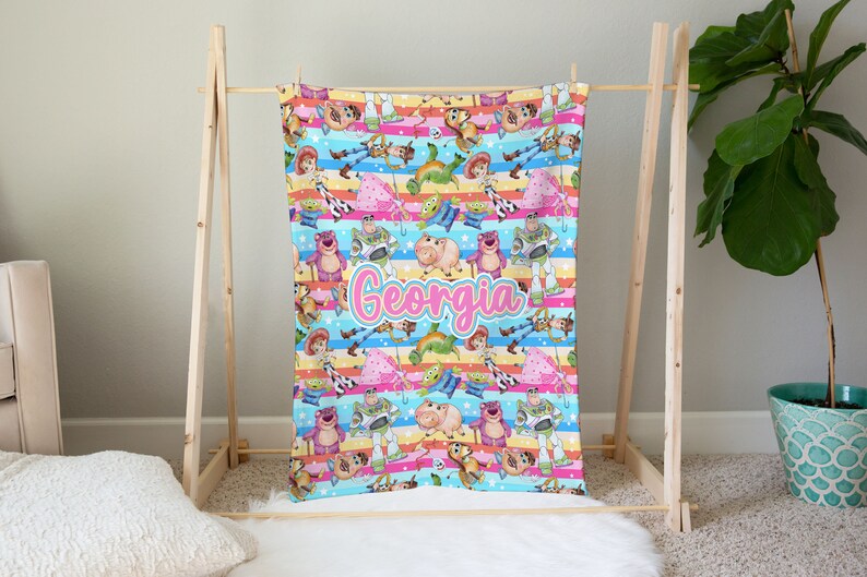 Peut inclure: Une couverture color&eacute;e avec un motif r&eacute;p&eacute;t&eacute; de personnages de la franchise de films Toy Story. La couverture est bleue, rose, jaune et verte avec des rayures blanches. Le nom "Georgia" est &eacute;crit en lettres cursives roses au centre de la couverture.