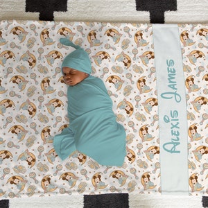 May include: A white baby blanket with a blue border and a repeating pattern of Mickey Mouse characters holding a rattle. The blanket has a personalized blue border with the text "Alexis James". A baby is swaddled in a blue swaddle and sleeping on the blanket.