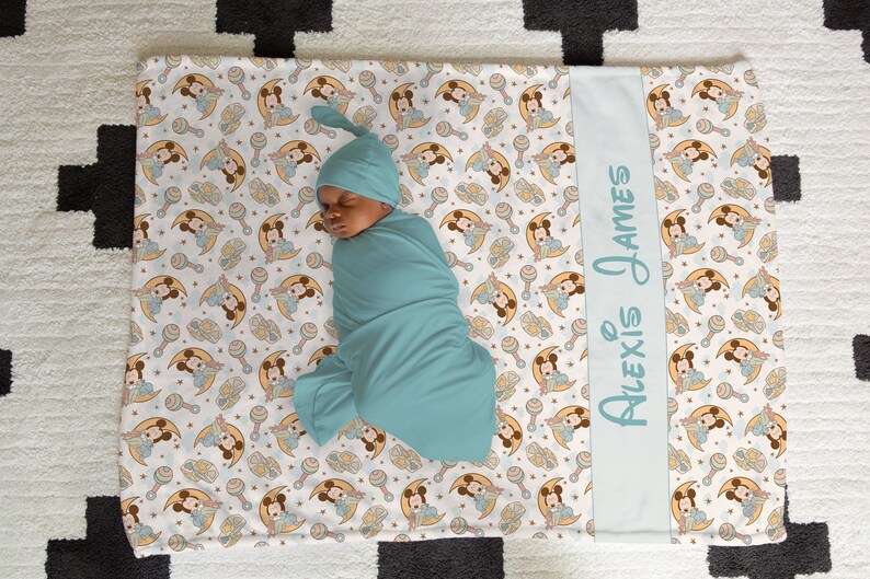 May include: A white baby blanket with a blue border and a repeating pattern of Mickey Mouse characters holding a rattle. The blanket has a personalized blue border with the text "Alexis James". A baby is swaddled in a blue swaddle and sleeping on the blanket.