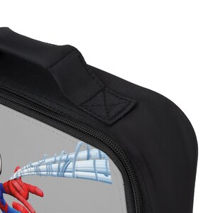 May include: A black lunch bag with a zipper closure and a padded handle. The bag features a Spiderman design with a gray background and a web design.