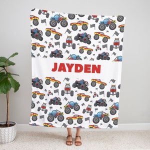 May include: White blanket with a red, blue, and yellow monster truck pattern. The blanket has the name "JAYDEN" printed in red letters in the center.