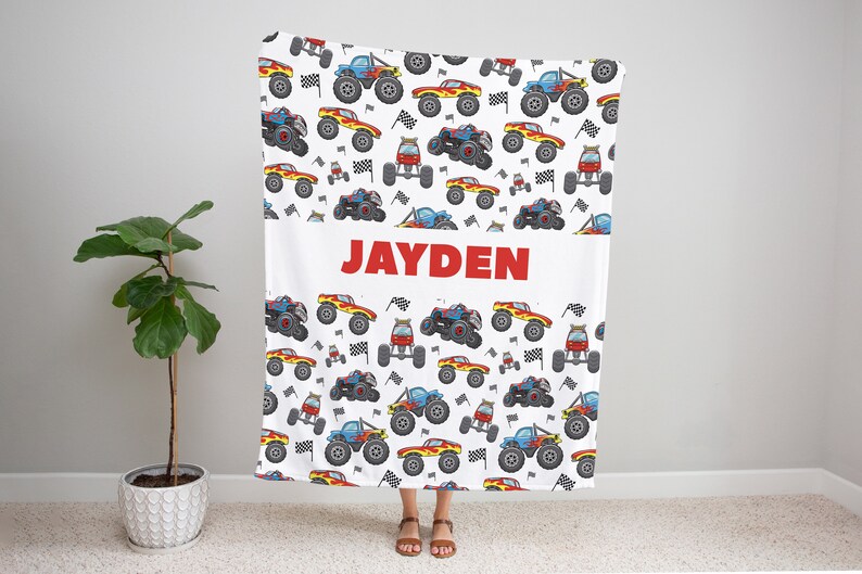 May include: White blanket with a red, blue, and yellow monster truck pattern. The blanket has the name "JAYDEN" printed in red letters in the center.