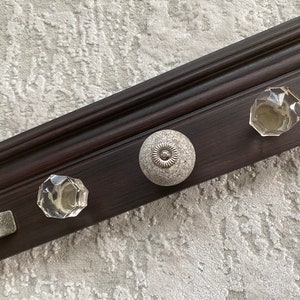 May include: A dark brown wooden shelf with three decorative knobs. Two of the knobs are clear glass and one is white with a textured surface.