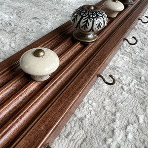May include: A wooden wall-mounted rack with four decorative ceramic knobs and four metal hooks. The rack is stained a dark brown color and has a grooved design.