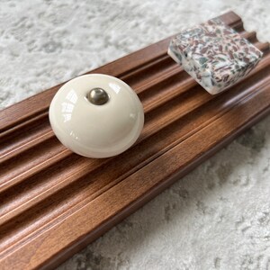 May include: Two cabinet knobs on a wooden surface. One knob is round and white with a silver accent. The other knob is square and has a brown, white, and green speckled pattern.