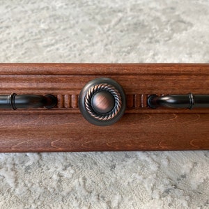 May include: A dark brown wooden shelf with bronze-colored hardware. The hardware includes two handles and a central circular knob with a rope-like design. The shelf has a decorative edge and is set against a neutral background.