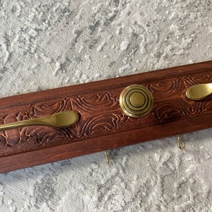May include: A wooden key holder with three hooks and two brass handles. The wood is dark brown and has a carved design. The handles are round and have a concentric circle design.