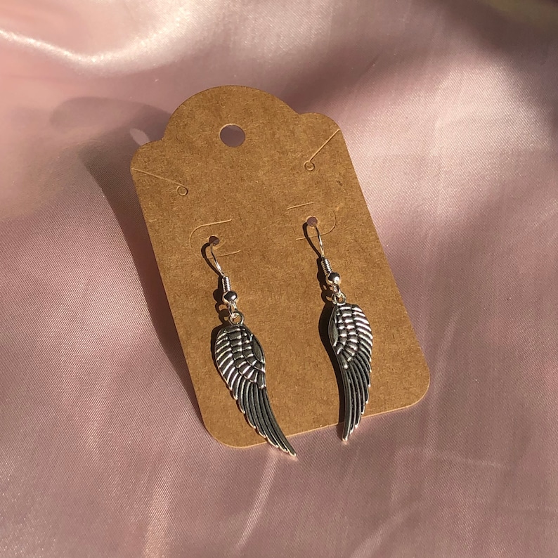 May include: A pair of silver angel wing earrings on a brown card. The wings are detailed and have a slightly textured finish.