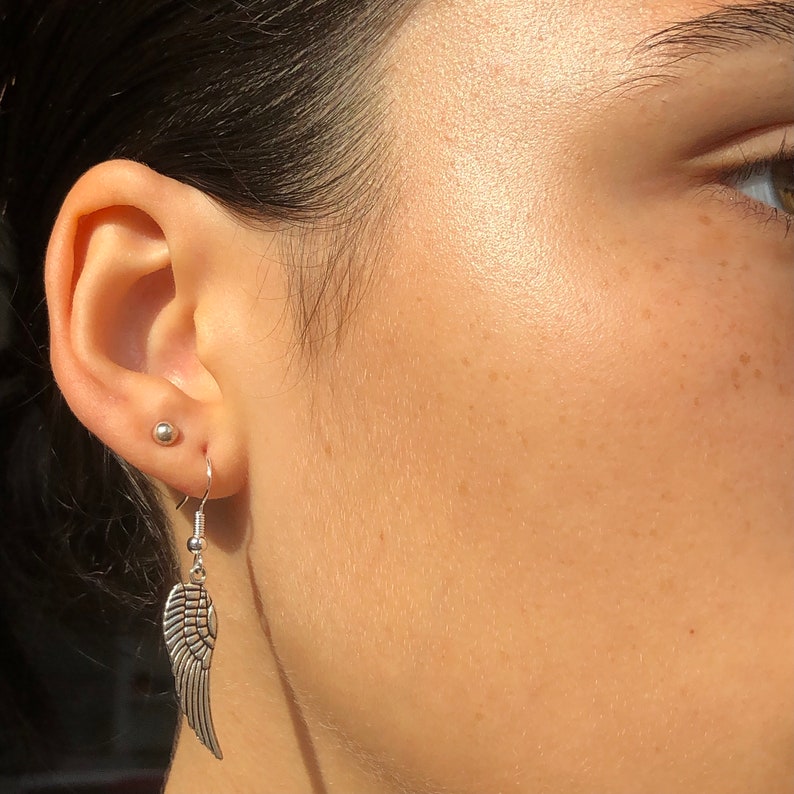 May include: Silver angel wing earrings with a hook closure. The earrings are worn in a person's ear.